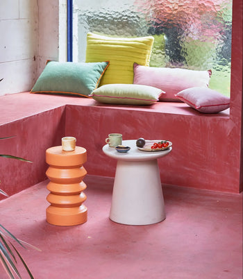 A pink seating nook with pastel cushions, an HKLIVING Stoneware Side Table by HKLIVING holding a cup and plate of food, and an orange sculptural stool; a large textured window lets in light.