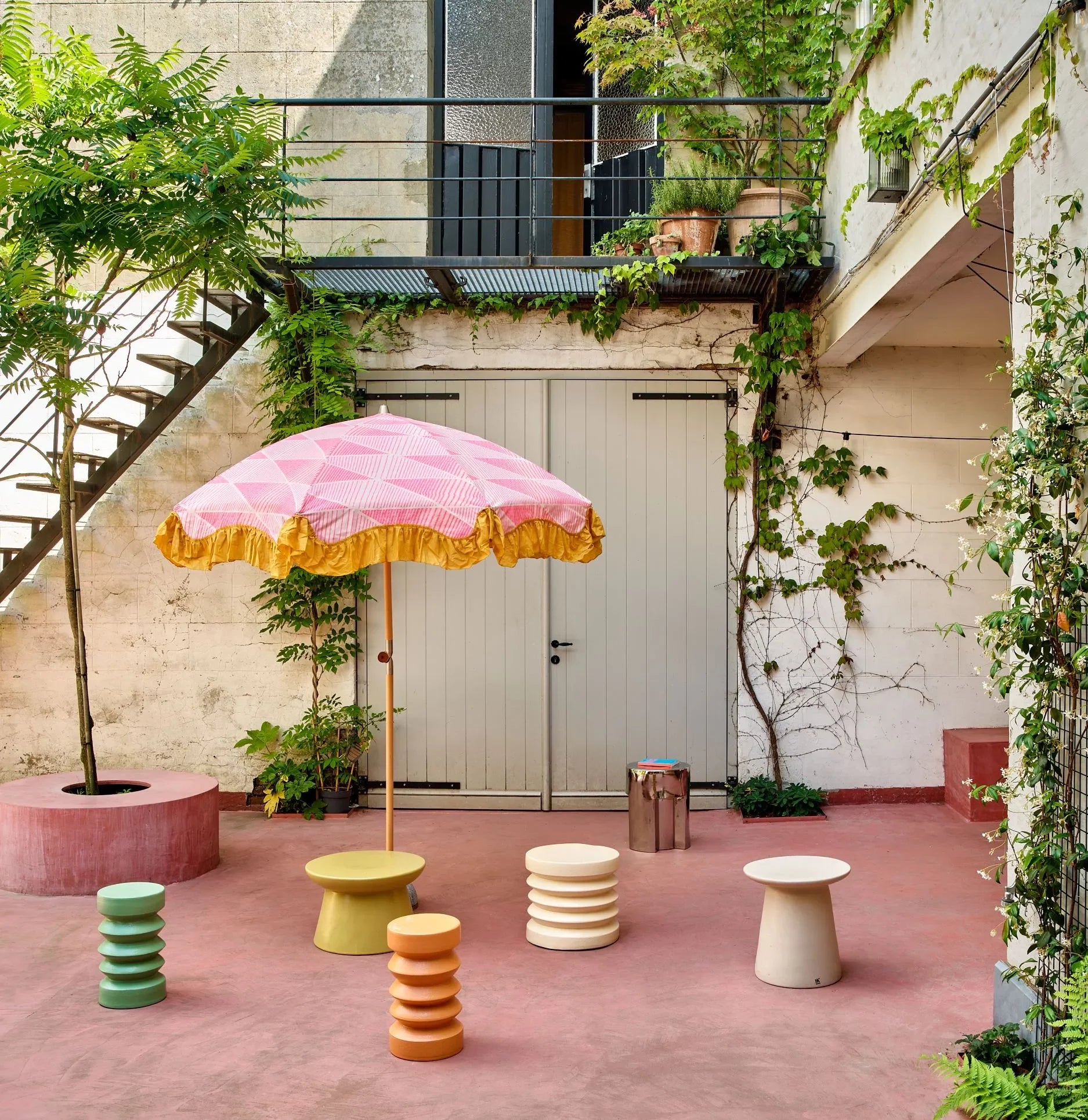 A vibrant patio features a pink and yellow umbrella, four pastel ribbed stools, the HKLIVING Stoneware Side Table by HKLIVING, a tree, potted plants, and wall greenery. A staircase leads to an upper balcony with lush plants.