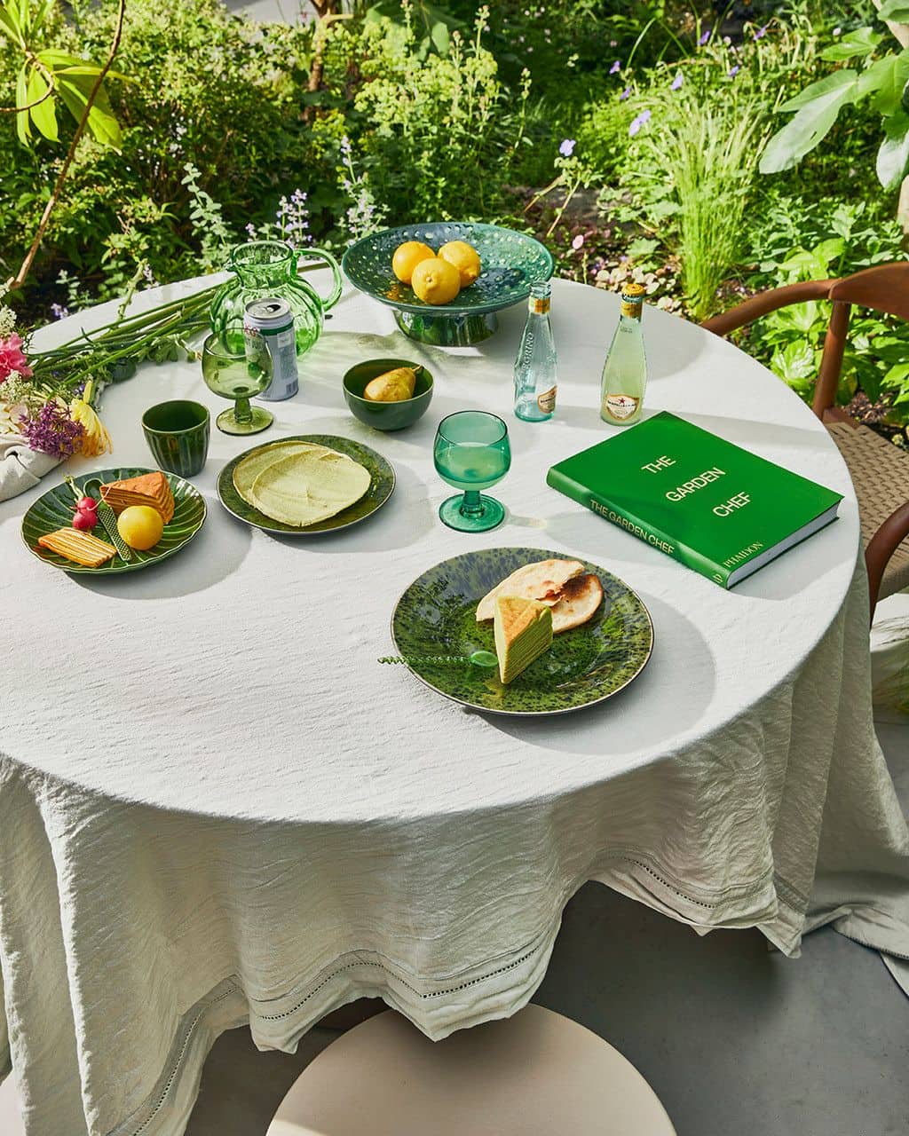A round table set outdoors with a white tablecloth features green dishware, HKliving The Emeralds: Ceramic Dessert Bowl (Set of 4), lemons, drinks, The Garden Chef book, and cake, surrounded by lush garden plants.