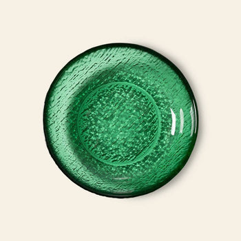 A round green glass plate with a textured pattern is shown from above on a light beige background, reflecting the modern elegance of HKliving’s The Emeralds: Glass Dessert Bowl collection.