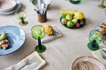 A dinner table set with HKliving The Emeralds: Glass Side Plates, green glass goblets, plates with figs, cutlery on napkins, and a fruit bowl of bananas, apples, and a lemon on a beige tablecloth.