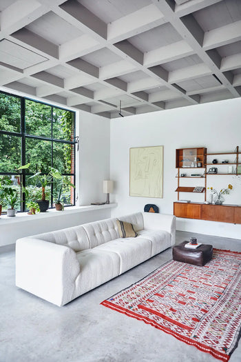 A modern living room features the roomy HKLIVING Vint Couch, Left, 1.5-Seat, a red patterned rug, brown ottoman, floating wooden shelf with decor, big window with plants, artwork on a white wall, and a striking geometric grid ceiling.