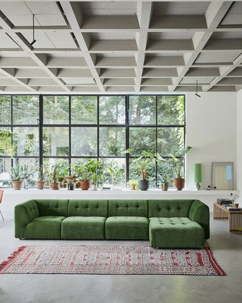 A modern living room with the HKLIVING Vint Couch, Left, 1.5-Seat in green velvet, a red and white patterned rug, potted plants on a white counter, large grid windows, a concrete coffered ceiling, and abundant natural light.