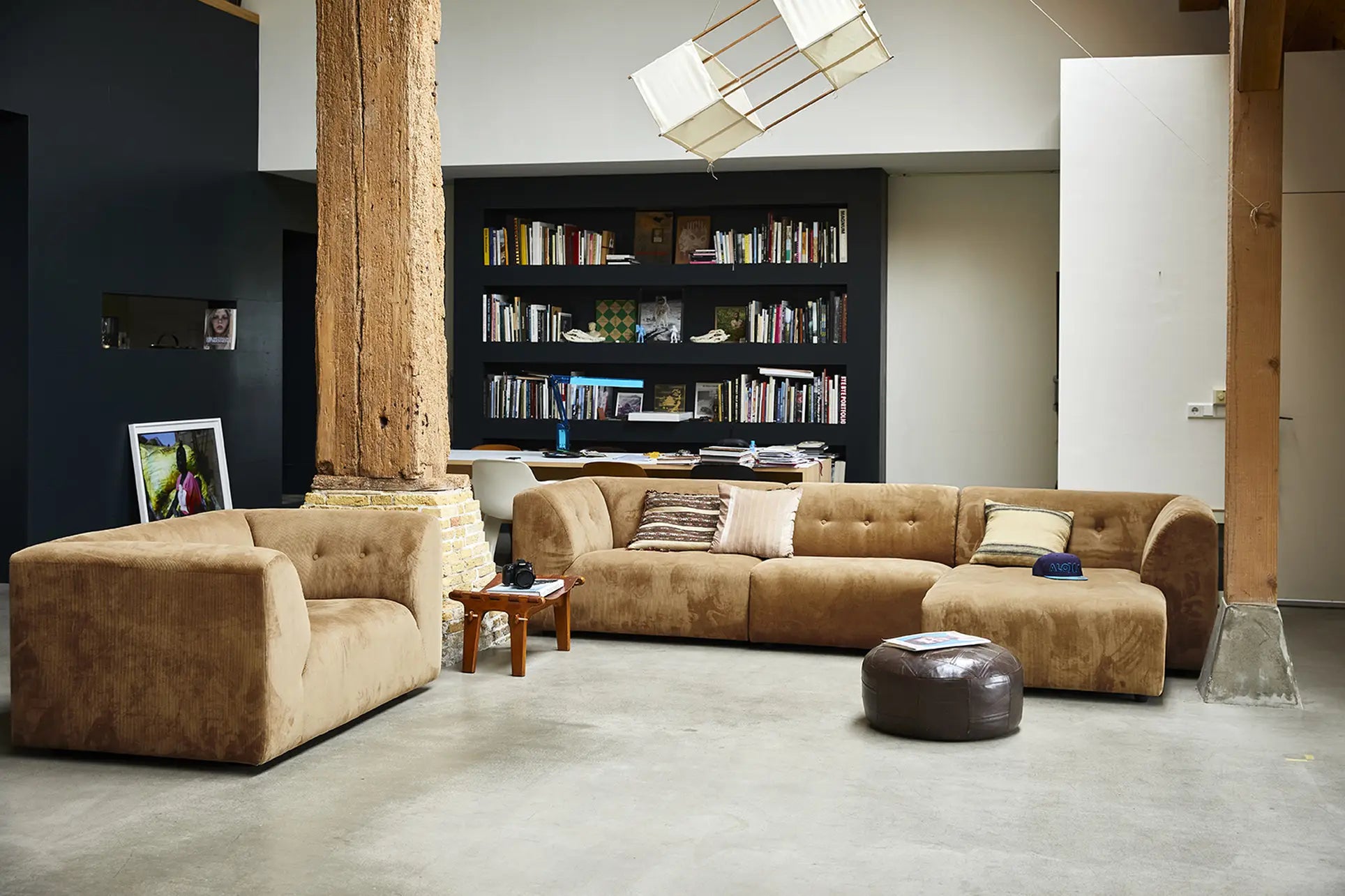 A modern living room with the HKLIVING Vint Couch Loveseat in brown suede, a wooden table, dark ottoman, exposed beams, and a large bookshelf against a black wall. The concrete floor and customizable sofa create an airy, bright space.