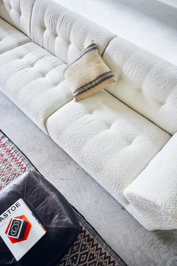A top-down view of the HKLIVING Vint Couch, Middle, 1.5-Seat in white with a beige pillow, beside a brown leather ottoman holding books and part of a patterned rug on a light floor.