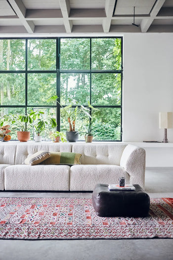 A modern living room with a large window features the HKLIVING Vint Couch, Middle, 1.5-Seat, a patterned rug, potted plants on the windowsill, a dark ottoman with books, a table lamp, and greenery visible outside.