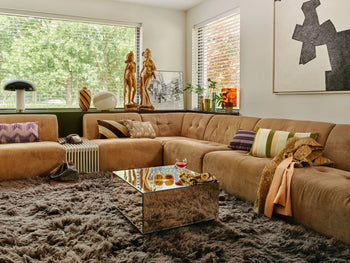 A cozy living room with the HKLIVING Vint Couch, Middle, 1.5-Seat modular sofa, striped pillows, a shaggy gray rug, mirrored coffee table with snacks and drink, large windows, sculptures, modern decor, and abstract wall art.