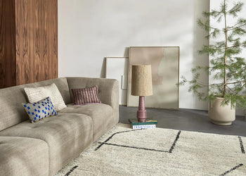 A modern living room featuring the HKLIVING Vint Couch, Middle, 1.5-Seat in beige, with patterned pillows, a white rug with black lines, a pink lamp on stacked books, two abstract artworks by the wall, and a potted plant in the corner.