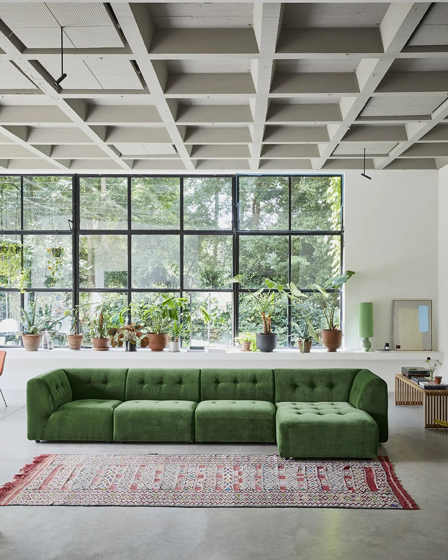 A modern living room featuring the HKLIVING Vint Couch, Right Divan, a patterned rug, and potted plants on a white ledge beneath tall grid windows that fill the space with natural light.