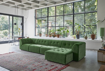 A modern living room features the HKLIVING Vint Couch, Right Divan in green, a red patterned rug, floor-to-ceiling windows, and potted plants on the windowsill that fill the space with natural light.