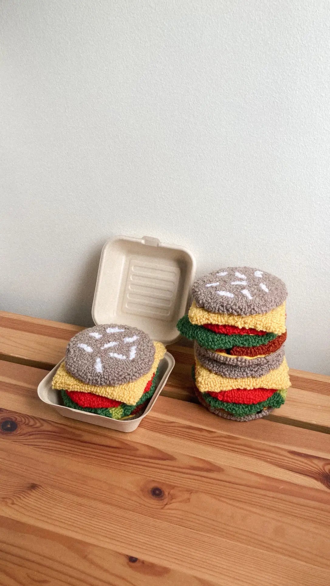 A set of Habichl Burger Coasters from Habichl, designed to resemble hamburgers, rests on a wooden surface beside a takeout container filled with more hamburger-shaped coasters.
