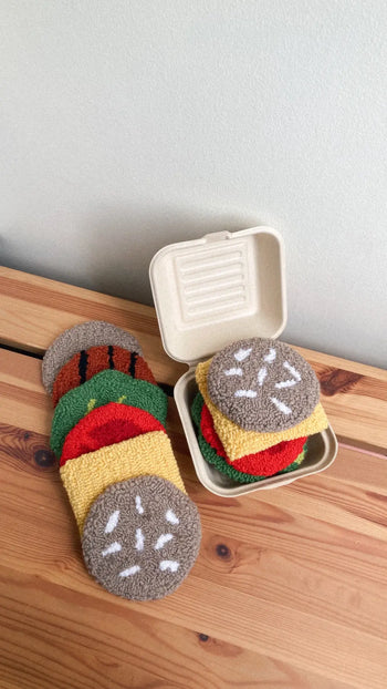 Several round, textured Habichl Burger Coasters from Habichl are stacked on a wooden table, with some inside an open, beige takeout container. These coasters resemble burger buns, lettuce, tomato, cheese, and patties.