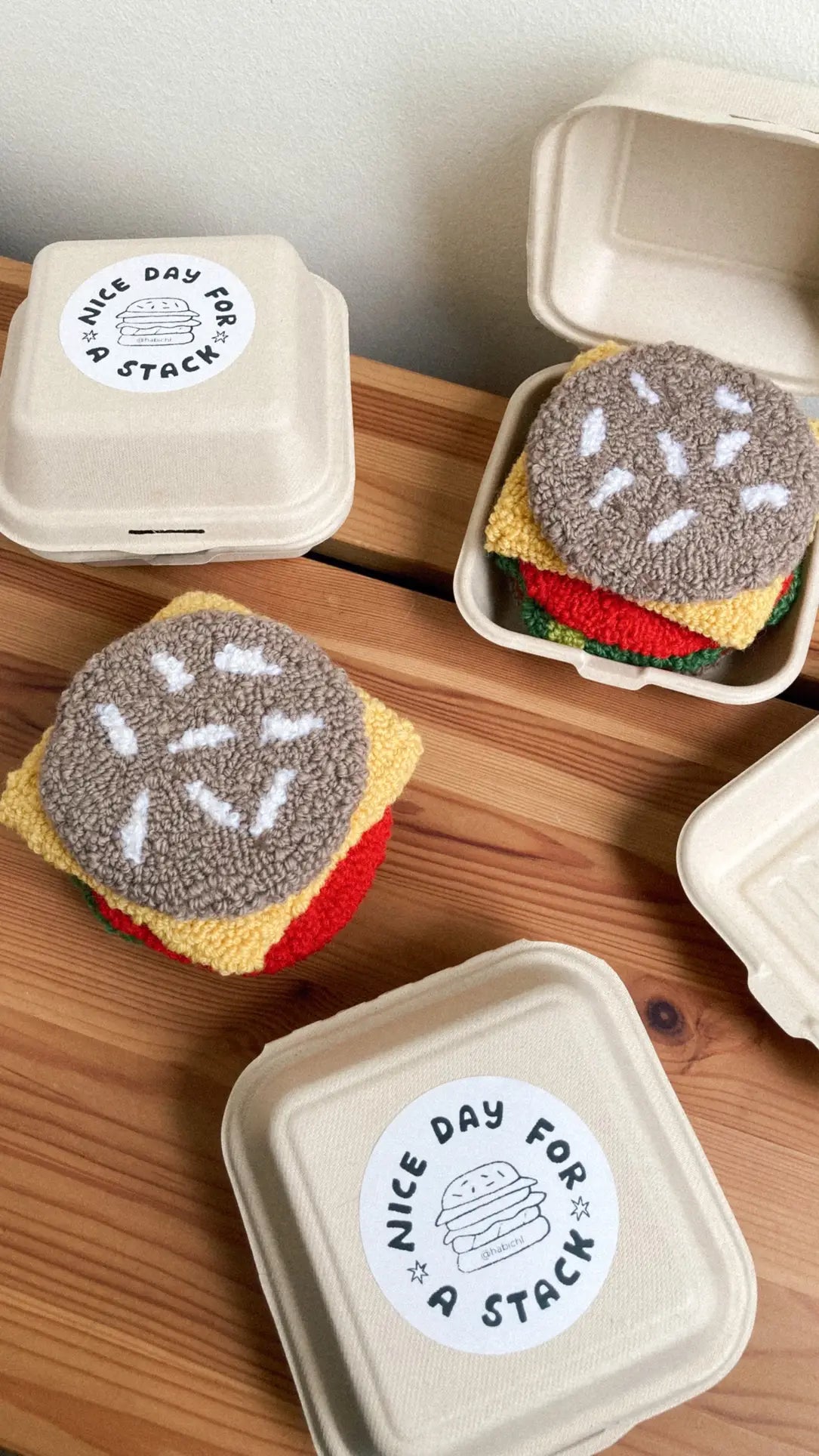 Three Habichl takeout boxes sit on a wooden table, each holding the Habichl Burger Coasters Set—cheeseburger-shaped coasters with sesame seed buns, topped with a “Nice Day For a Stack” sticker and playful burger illustration.