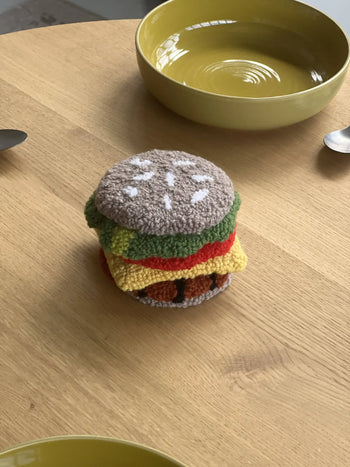 The Habichl Burger Coasters Set by Habichl, designed to look like a hamburger with layers of lettuce, tomato, cheese, and a sesame seed bun, sits on a wooden table between two green bowls and spoons.