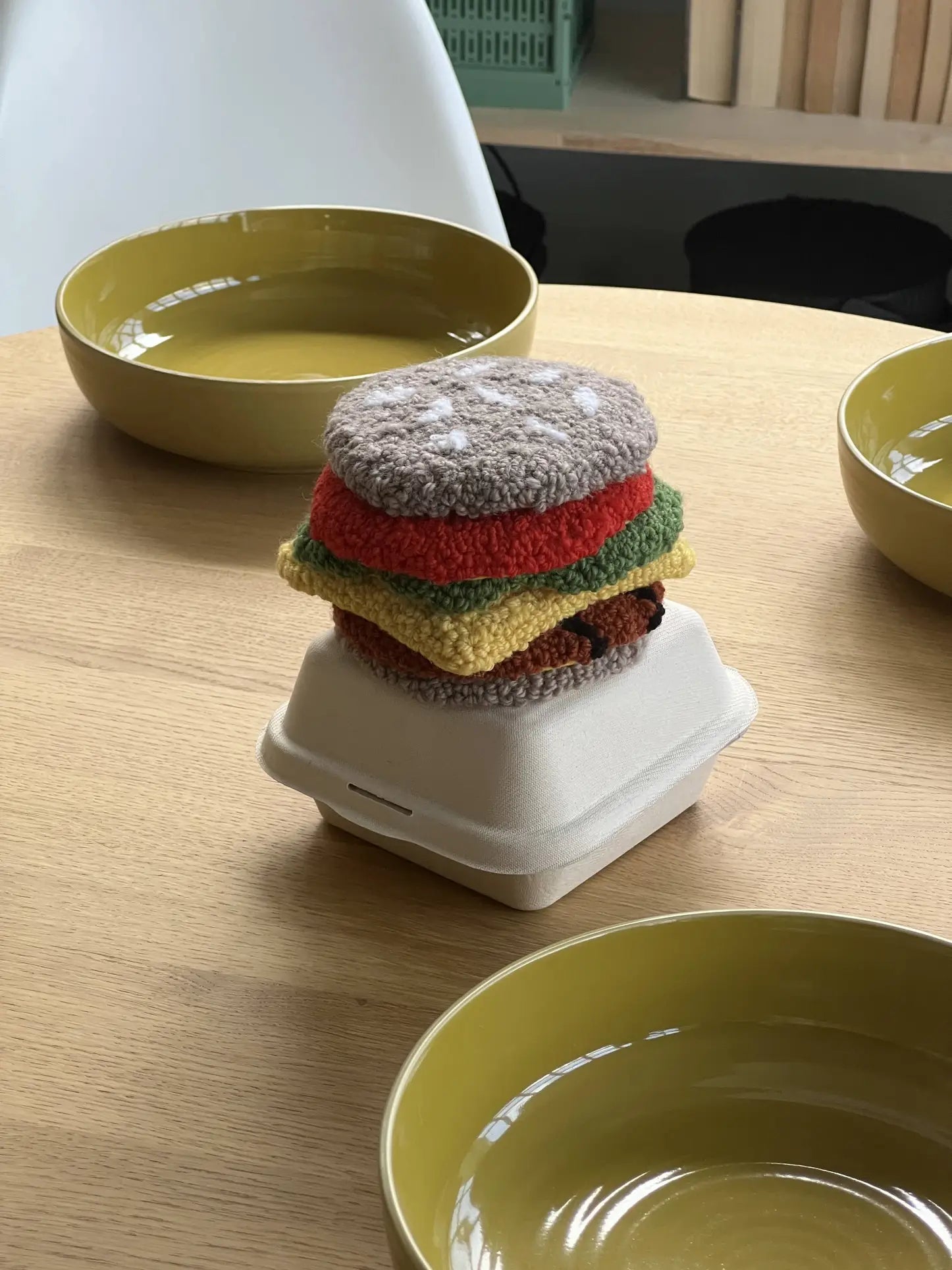 The Habichl Burger Coasters Set by Habichl, styled like a layered burger with bun, lettuce, tomato, cheese, and patty pieces, rests atop an open takeout box on a wooden table next to three empty yellow bowls.