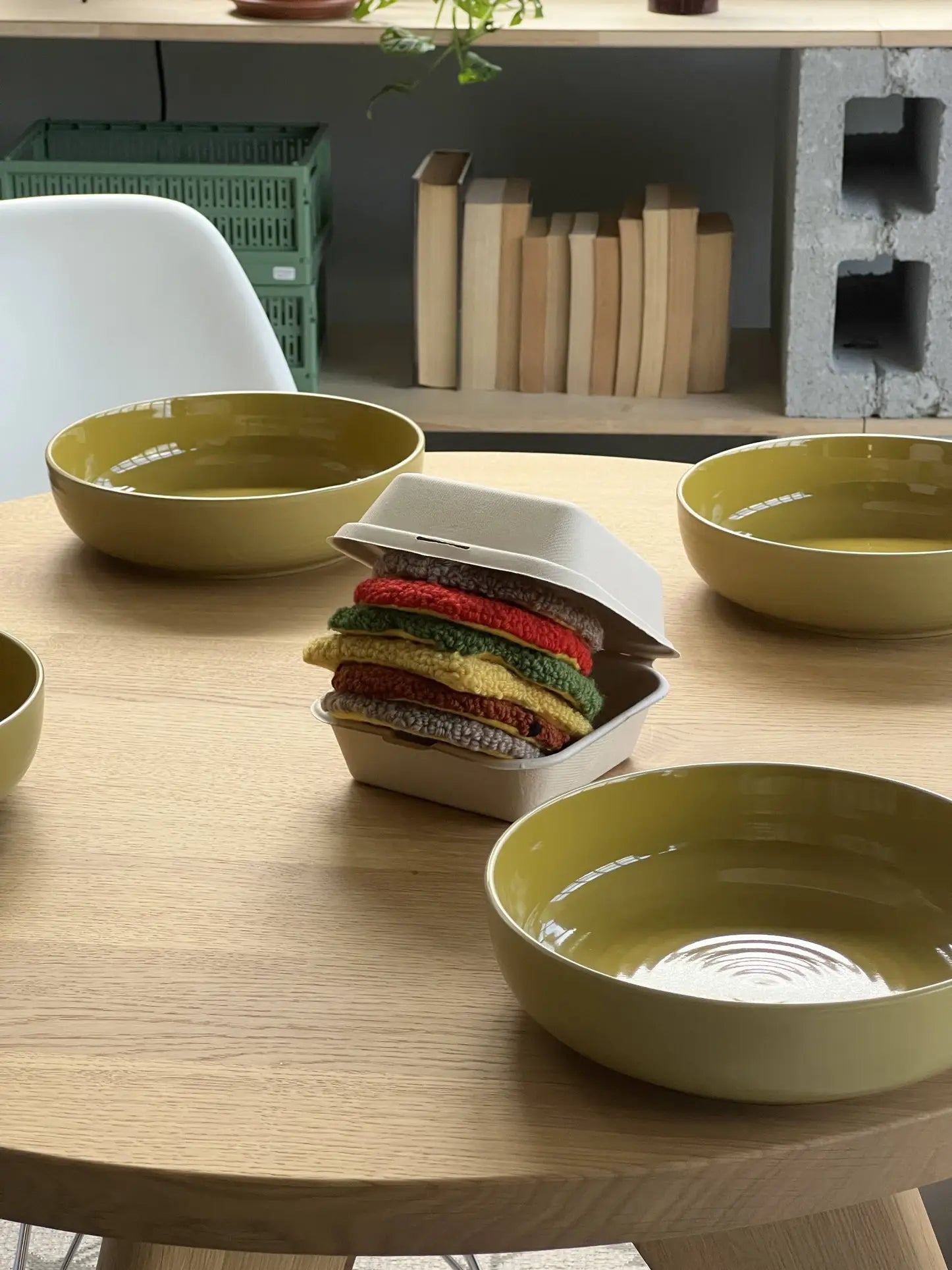 Four yellow bowls on a wooden table surround a takeout box from The Stacked Store, which holds the Habichl Burger Coasters Set by Habichl—colorful knitted coasters resembling a stacked burger. Books and crates sit on shelves behind.