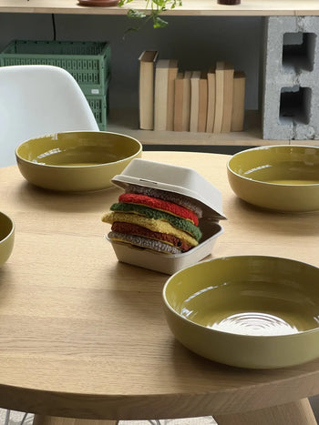 Four yellow bowls on a wooden table surround a takeout box from The Stacked Store, which holds the Habichl Burger Coasters Set by Habichl—colorful knitted coasters resembling a stacked burger. Books and crates sit on shelves behind.