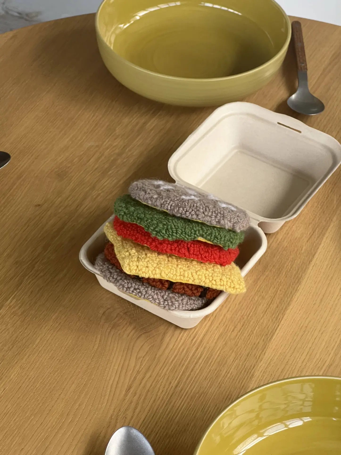 A takeout box from Habichl sits on a wooden table, holding the Habichl Burger Coasters Set—crocheted coasters stacked with bun, lettuce, cheese, tomato, and patty. Plates and utensils add a playful touch nearby.