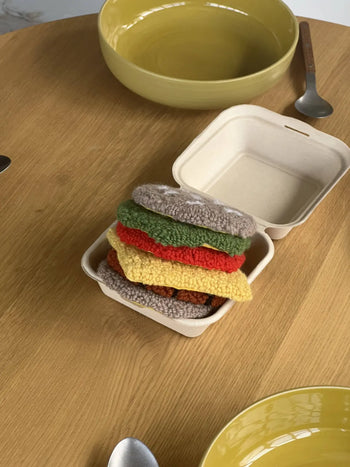 A takeout box from Habichl sits on a wooden table, holding the Habichl Burger Coasters Set—crocheted coasters stacked with bun, lettuce, cheese, tomato, and patty. Plates and utensils add a playful touch nearby.