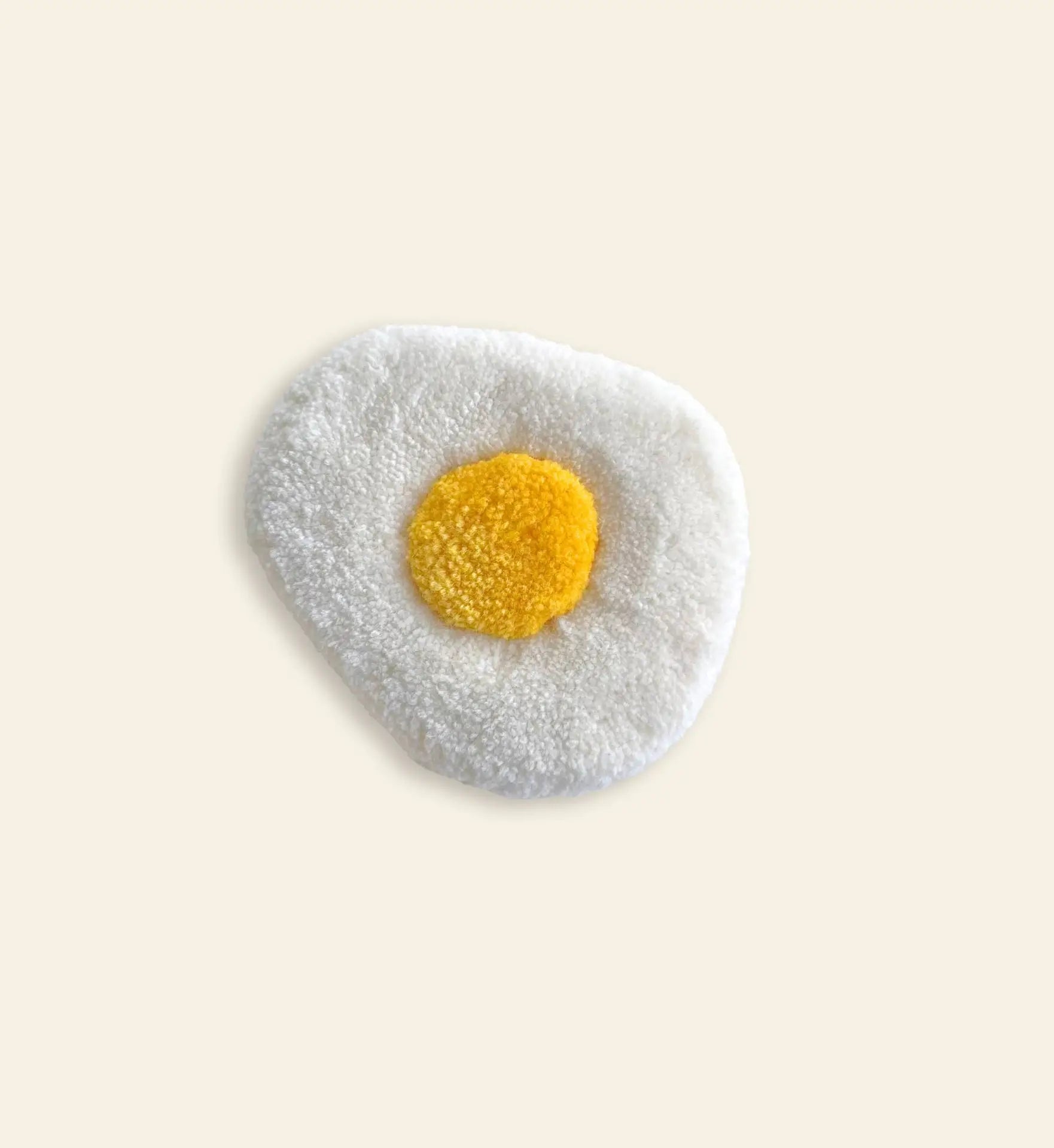 The Habichl Tufted Egg Coaster by Habichl features a textured white outer area and a yellow center, resembling a sunny side up egg, shown on a light beige background.