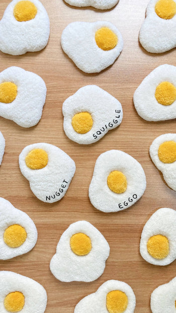 Habichl Tufted Egg Coasters by Habichl, shaped like sunny-side-up eggs, are displayed on wood. Names like NUGGET, SQUIGGLE, and EGGO appear on the whites, giving these playful coasters a handcrafted, plush toy look.