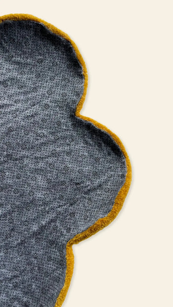 A close-up of a gray, flower-shaped fabric with a yellow border, set on a beige background. Only part of the whimsical Habichl Tufted Flower Floor Rug by Habichl is visible in the image.