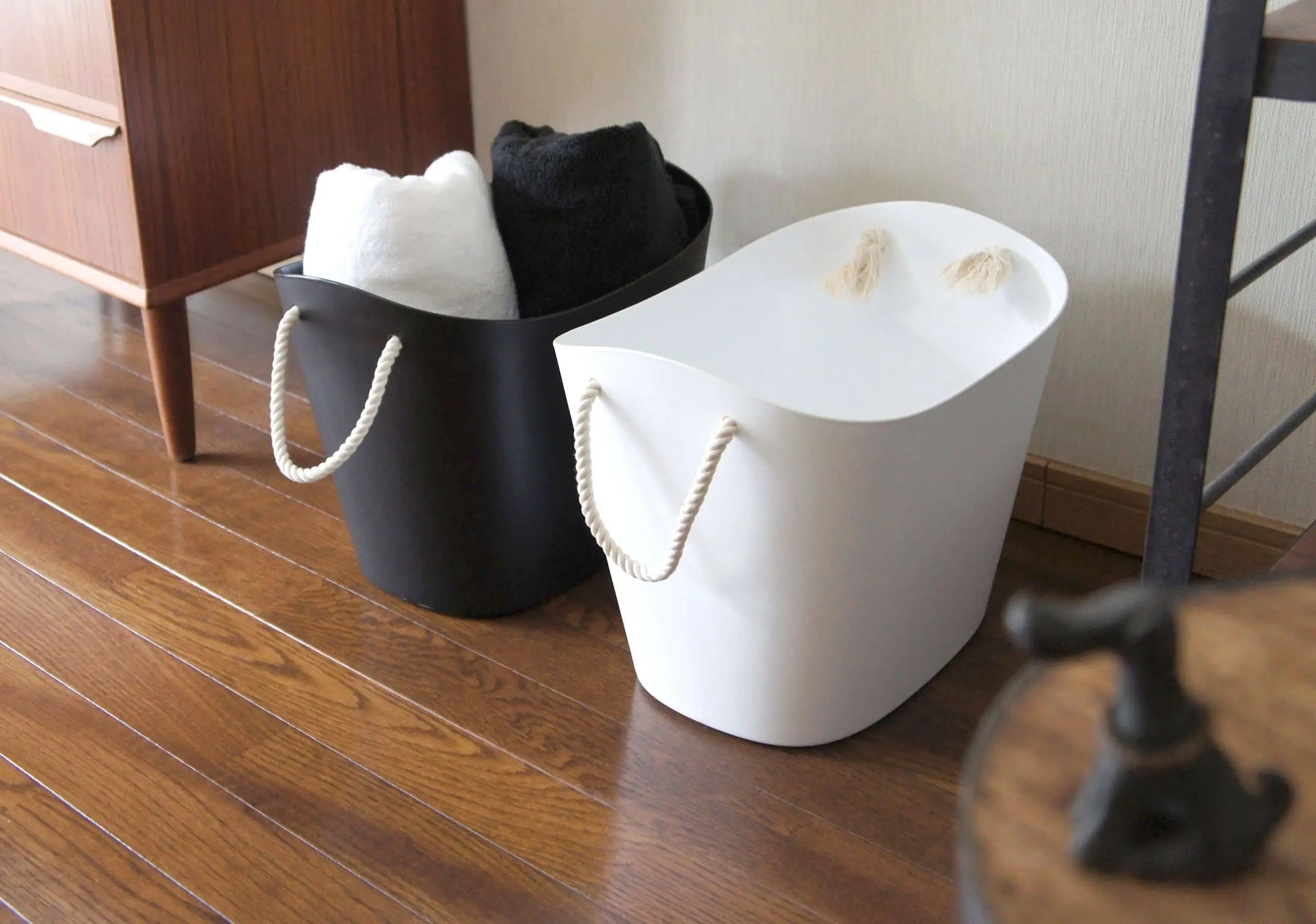 Two Hachiman Balcolore Storage Basket M bins—one black, one white—rest on a wooden floor. Rolled towels fill these minimalist Japanese baskets by Hachiman, providing stylish storage beside a piece of furniture.