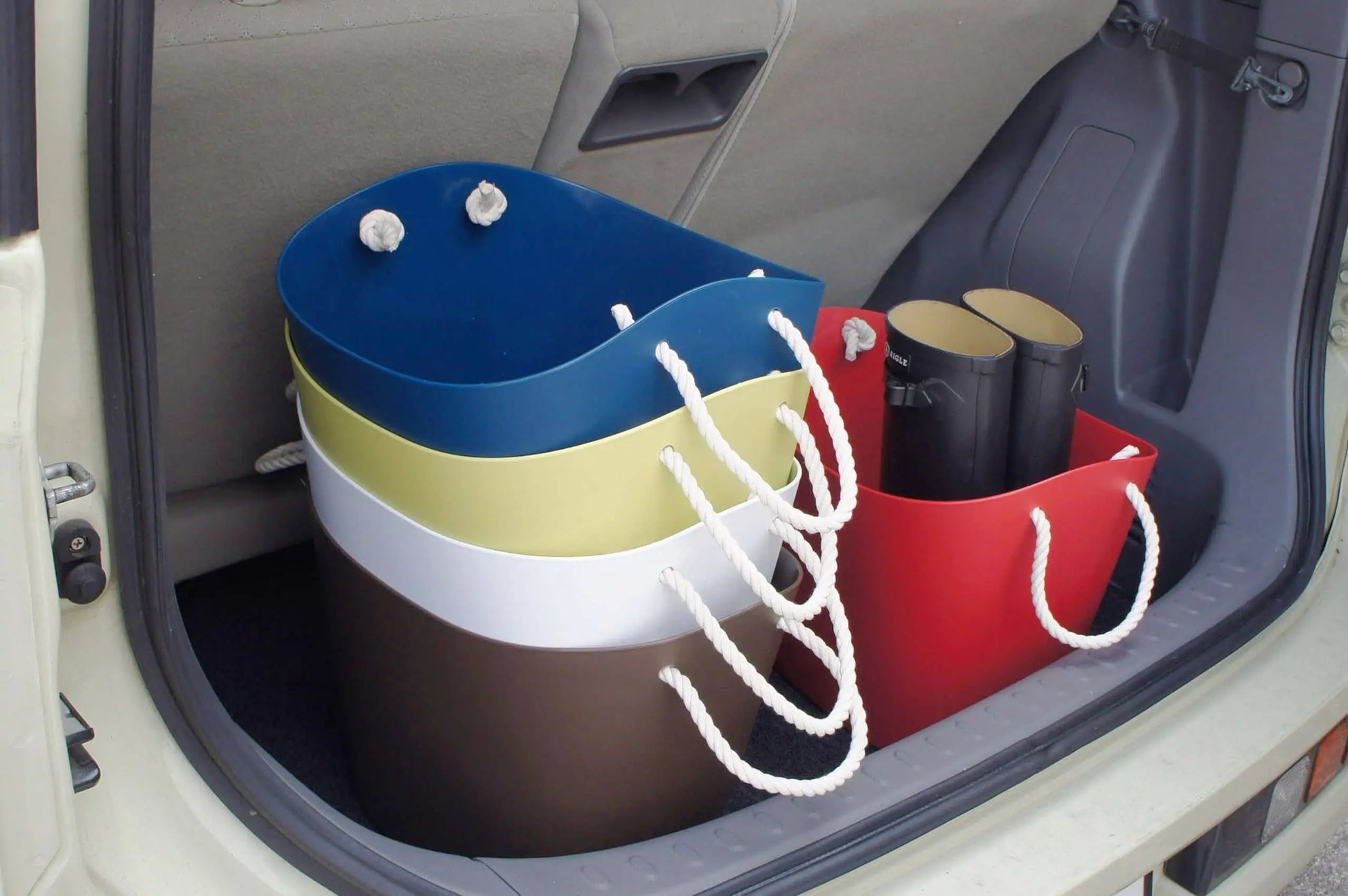 Four vibrant Hachiman Balcolore Storage Basket M bins with rope handles and two rolled-up mats are neatly arranged in a car trunk. The Japanese-designed baskets are nested together, with the mats stored inside a red basket for versatile use.