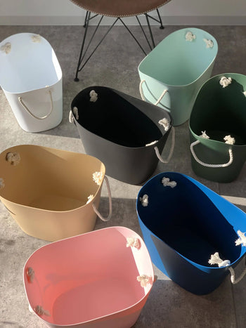 Seven Hachiman Balcolore Storage Basket M tubs in white, black, green, tan, blue, pink, and mint are arranged in a semi-circle on a gray floor, showing off their minimalist Japanese style and versatile rope handles.
