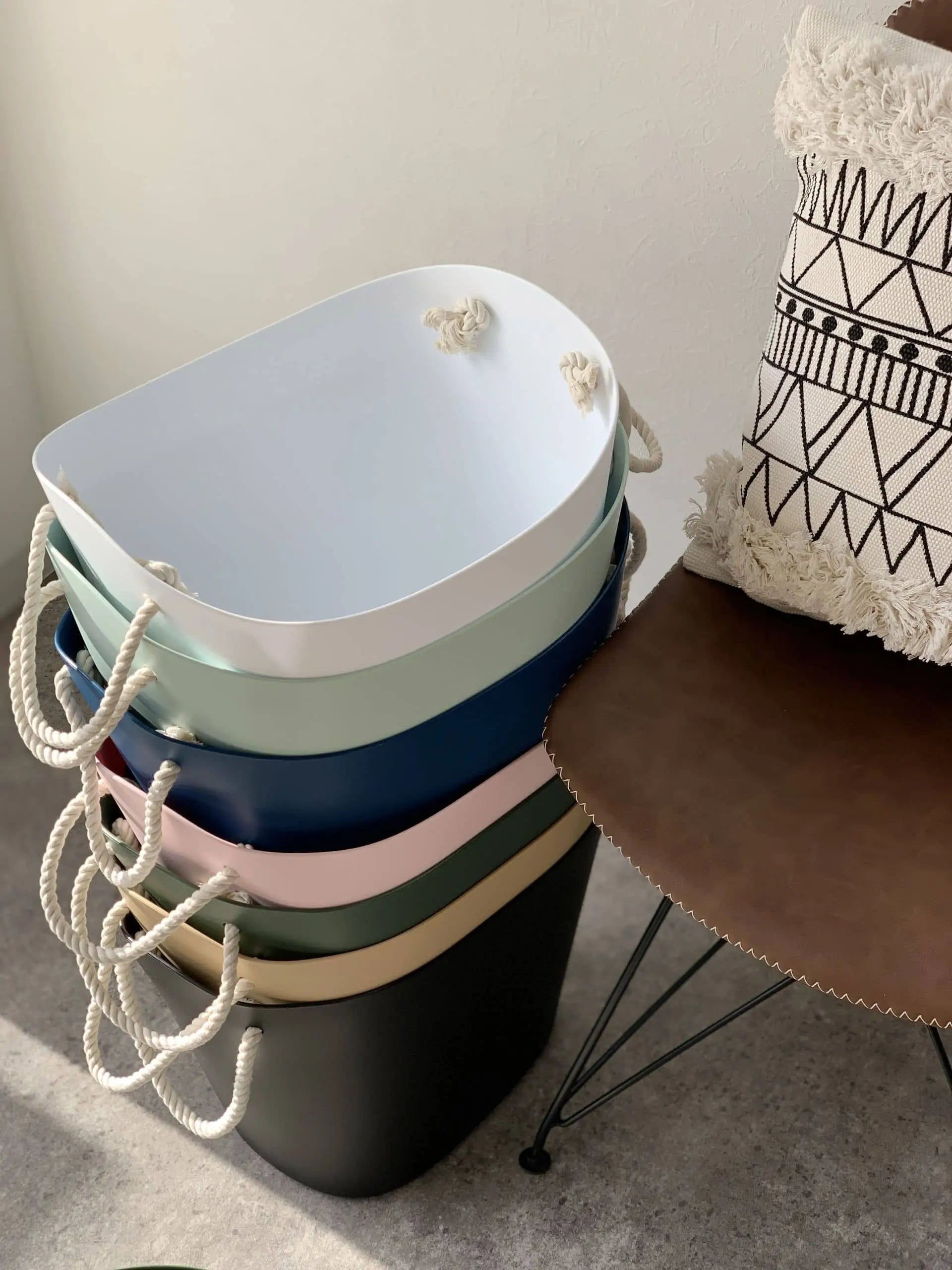 A stack of Hachiman Balcolore Storage Basket M organizers with rope handles sits beside a brown chair with a geometric cushion, bringing minimalist Japanese style and versatile storage to the bright room.