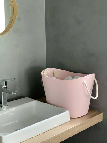 A pink Hachiman Balcolore Storage Basket M with rope handles sits on a wooden countertop by a white sink in a gray-walled, minimalist Japanese bathroom—ideal for stylish, multi-purpose storage.