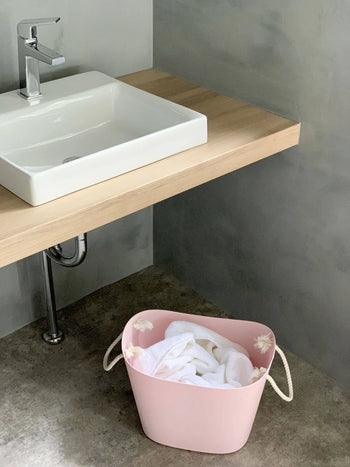 A Hachiman Balcolore Storage Basket M in pink with white handles, filled with white clothes, is placed on the floor under a wooden countertop with a modern white sink and silver faucet in a minimalist Japanese bathroom.