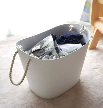 A Hachiman Balcolore Storage Basket M with rope handles, filled with folded clothes, rests on a light carpet near a window and wooden furniture—perfect for minimalist Japanese or versatile storage by Hachiman.