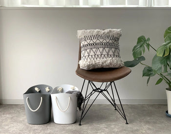 A brown chair with a patterned pillow sits beside two minimalist Japanese baskets—one gray and one white Hachiman Balcolore Storage Basket M—on a gray floor. To the right is a green potted plant, with a white curtain in the background.