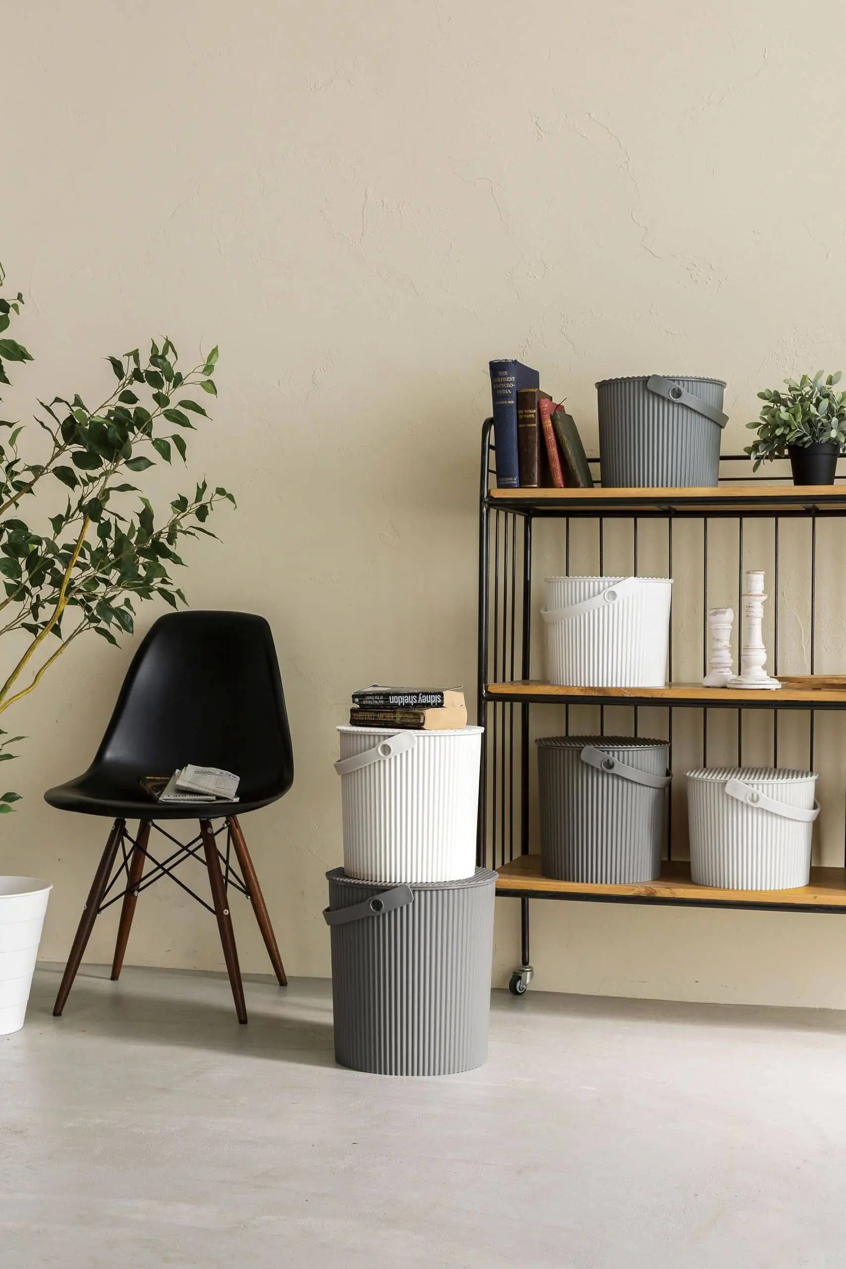 A minimalist room features a black chair, potted plants, and a black metal shelf with Hachiman Omnioutil Storage Bucket L by Hachiman, books, and decor—stylish storage perfect for indoor or outdoor use against a light beige wall.