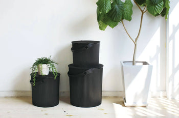 Three black Hachiman Omnioutil Storage Bucket S containers with handles are stacked beside a tall potted plant. One bucket holds a small trailing plant. Ideal for indoor or outdoor use, sunlight streams in from the right.