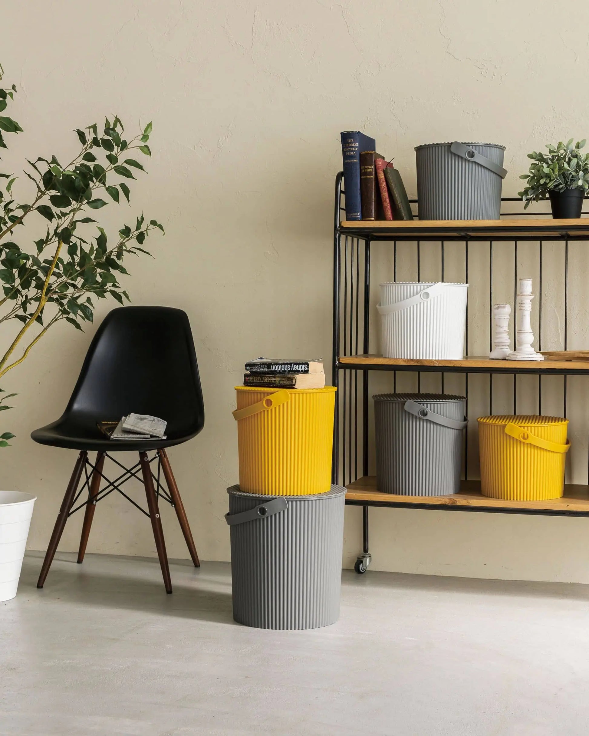 A modern room with a black chair, potted plant, and a shelf displaying Hachiman Omnioutil Storage Bucket S in yellow, gray, and white—ideal for indoor or outdoor use. Three more Hachiman buckets are stacked nearby for extra organization.
