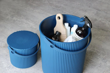 Two ribbed blue Hachiman Omnioutil Storage Bucket S bins sit on a gray floor—one holds cleaning supplies with the lid off, the other is closed. Durable and versatile, Hachiman buckets are ideal for indoor or outdoor storage needs.
