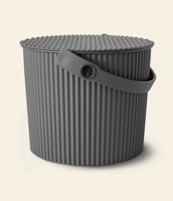 The Hachiman Omnioutil Storage Bucket S by Hachiman is a round, dark gray bucket with a ribbed lid and flexible handle, ideal for indoor or outdoor use. Its shown against a plain light background.