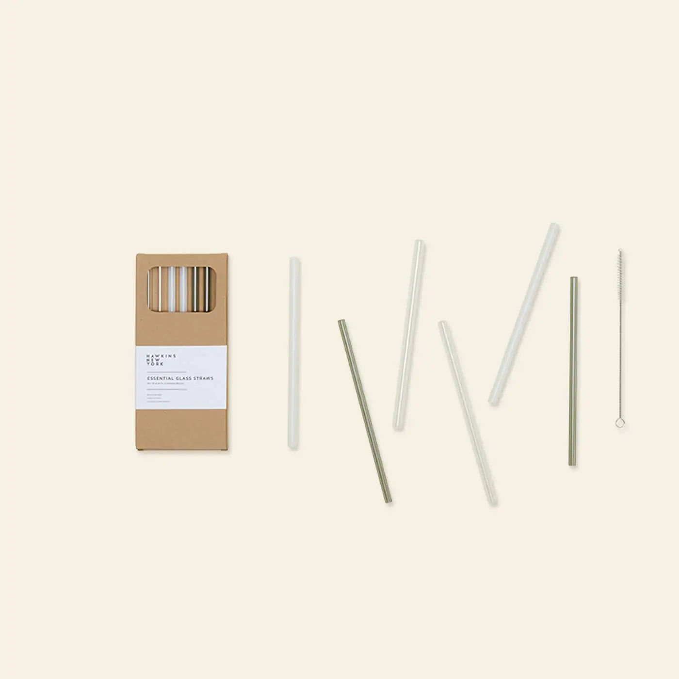 The Hawkins New York Essential Glass Straws, Set of 6, features reusable borosilicate glass straws in assorted colors with a cleaning brush, displayed beside their cardboard packaging on a light beige background.