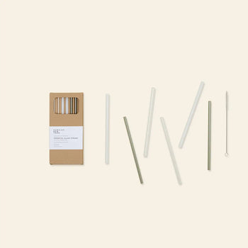 The Hawkins New York Essential Glass Straws, Set of 6, features reusable borosilicate glass straws in assorted colors with a cleaning brush, displayed beside their cardboard packaging on a light beige background.