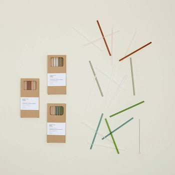 Three Hawkins New York Essential Glass Straws, Set Of 6 boxes are displayed on a light surface, with assorted colored reusable glass straws and a cleaning brush from Hawkins New York arranged nearby.