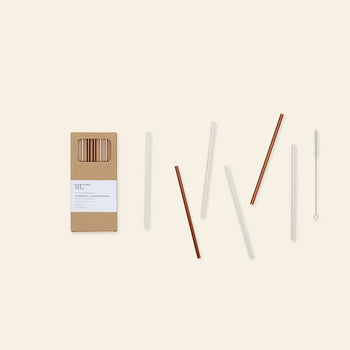 The Hawkins New York Essential Glass Straws Set includes six reusable borosilicate glass straws (three clear, three brown) and a cleaning brush, all from Hawkins New York, displayed on a light beige background.