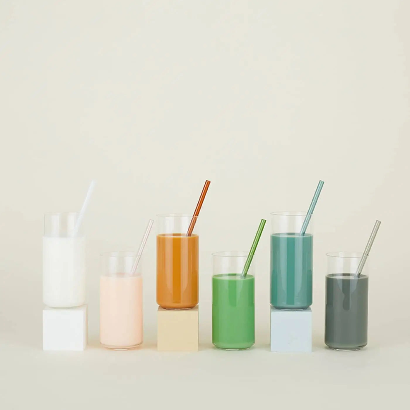 Six colorful drinks, each with a matching straw from the Hawkins New York Essential Glass Straws Set of 6, are displayed in a row on blocks against a neutral background.