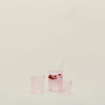 Three translucent pink Hawkins New York Essential Glassware, Set Of 4, Medium glasses sit on a plain light surface. The tallest holds ice, a clear straw, and pink liquid; the other two remain empty against a neutral background.