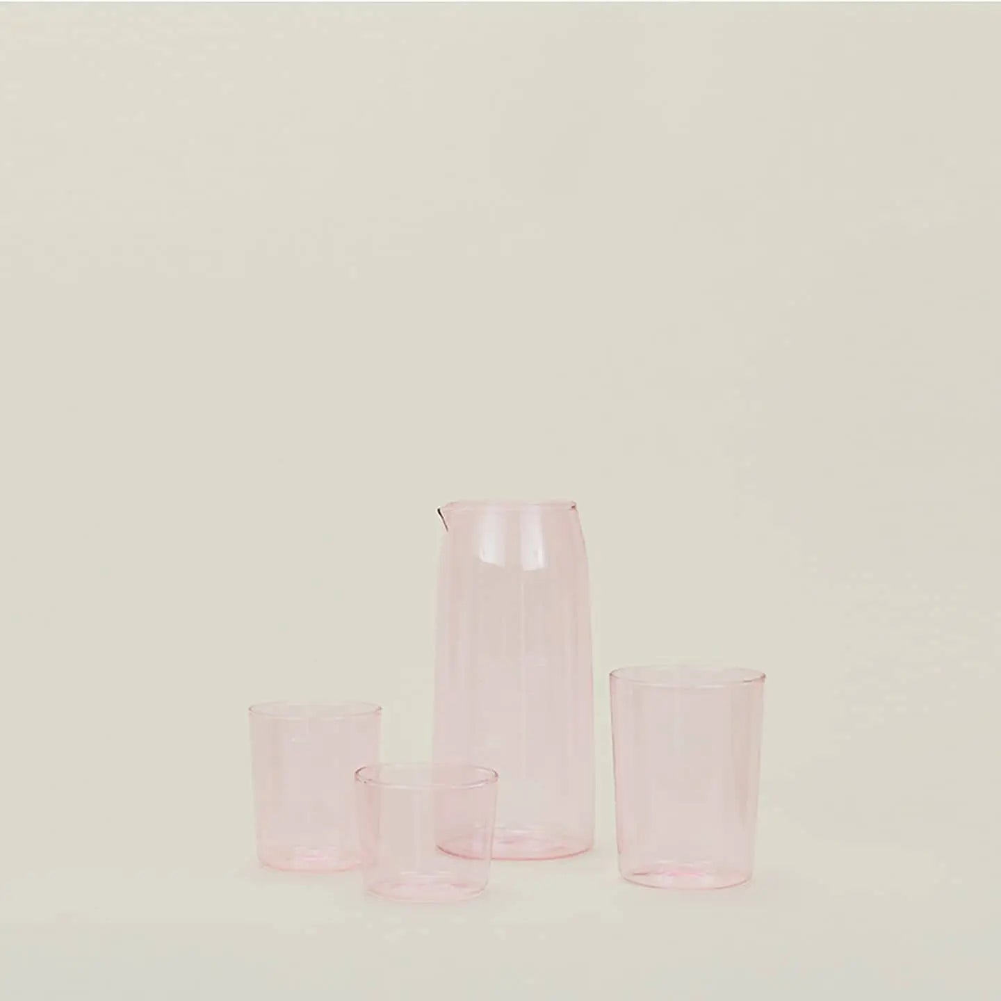 The Hawkins New York Essential Glassware Set Of 4 (Medium) by Hawkins New York features translucent pink glassware with a tall pitcher and three matching glasses of varying heights, crafted from premium glass and set against a plain light background.
