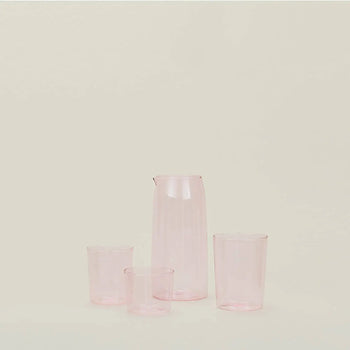 The Hawkins New York Essential Glassware Set Of 4 (Medium) by Hawkins New York features translucent pink glassware with a tall pitcher and three matching glasses of varying heights, crafted from premium glass and set against a plain light background.