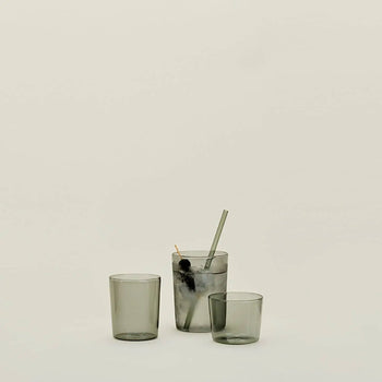 Three Hawkins New York Essential Glassware medium drinking glasses sit on a plain light background; one, highlighting quality glass construction, holds ice, a dark cherry, a metal straw, and cocktail pick while the other two remain empty.