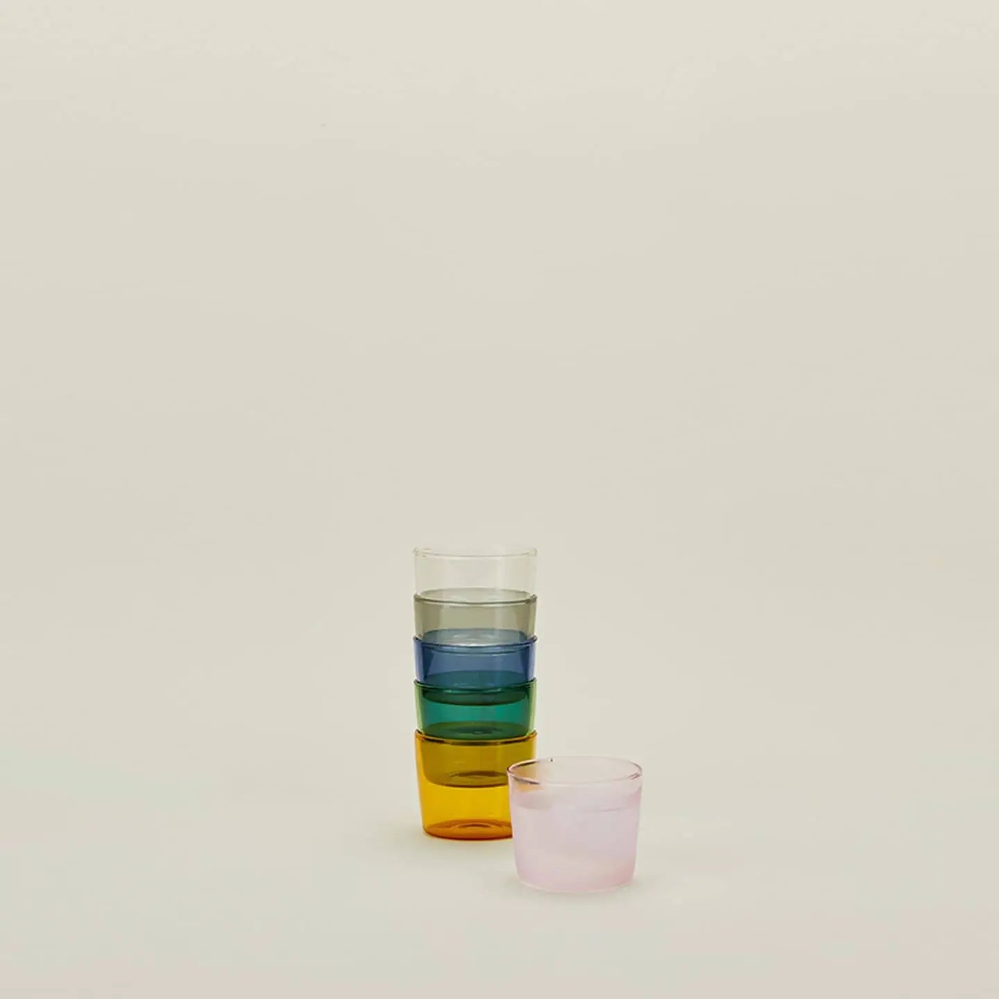 Four colorful glass cups from the Hawkins New York Essential Glassware Set of 4 (Small) are stacked on a beige background, with another cup placed separately in front.