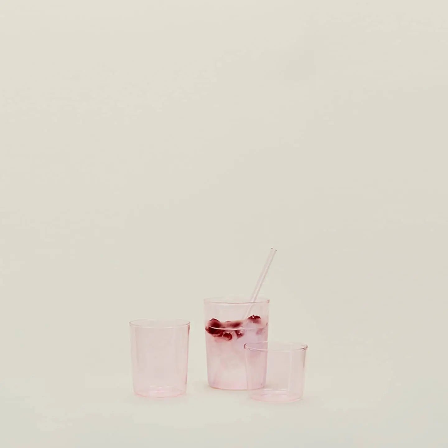 Three translucent pink Hawkins New York Essential Glassware small glasses are arranged on a light surface—one with ice, a straw, and a red garnish; two are empty. The background is plain and off-white. Set of 4 by Hawkins New York.